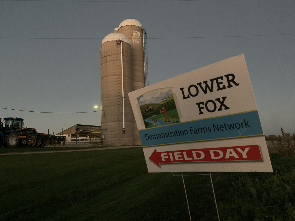 A yard sign in the foreground says "Lower Fox Demonstration Farms Network Field Day". In the background is a farm at dusk with a tractor and two large grain silos.