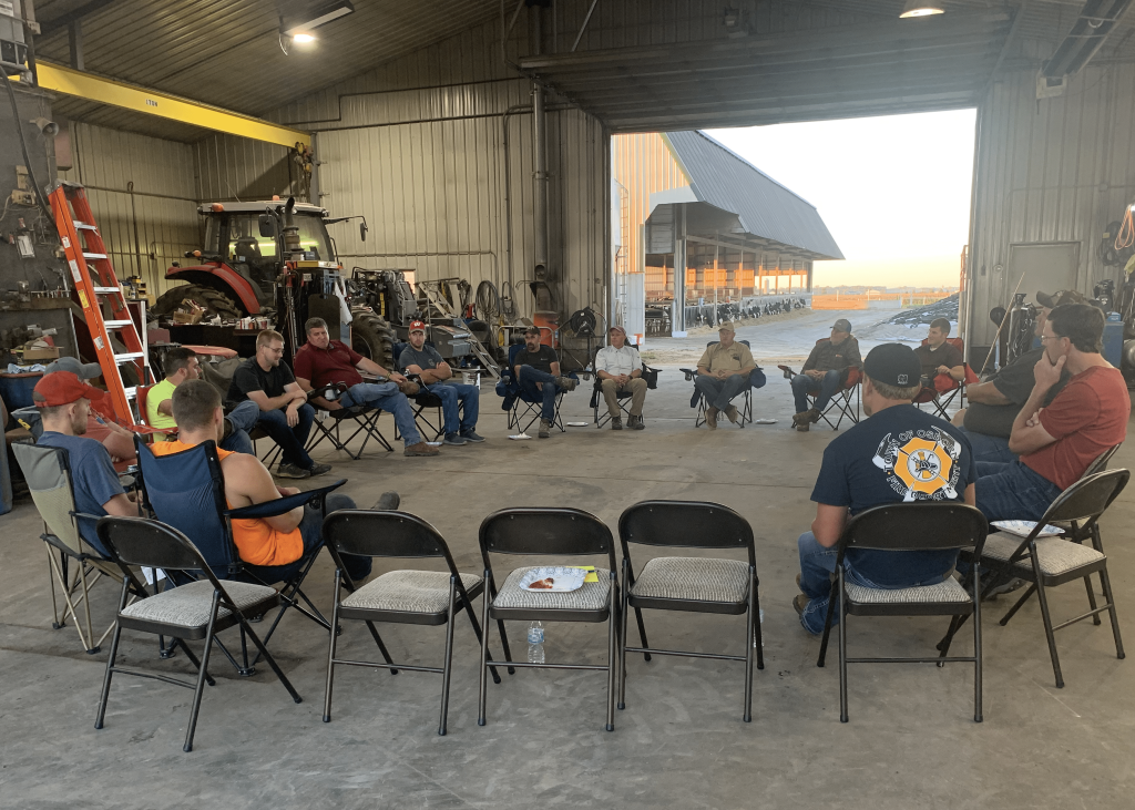 A group of people sit in a circle of chairs in a large metal barn.