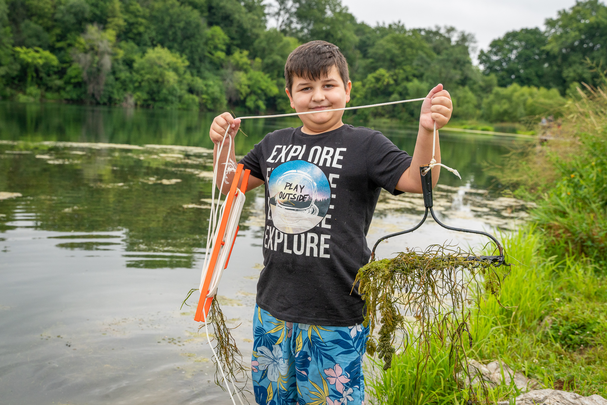 A young boy standing on the bank of a small stream holding a rake head tied to a string with aquatic weeds in it.