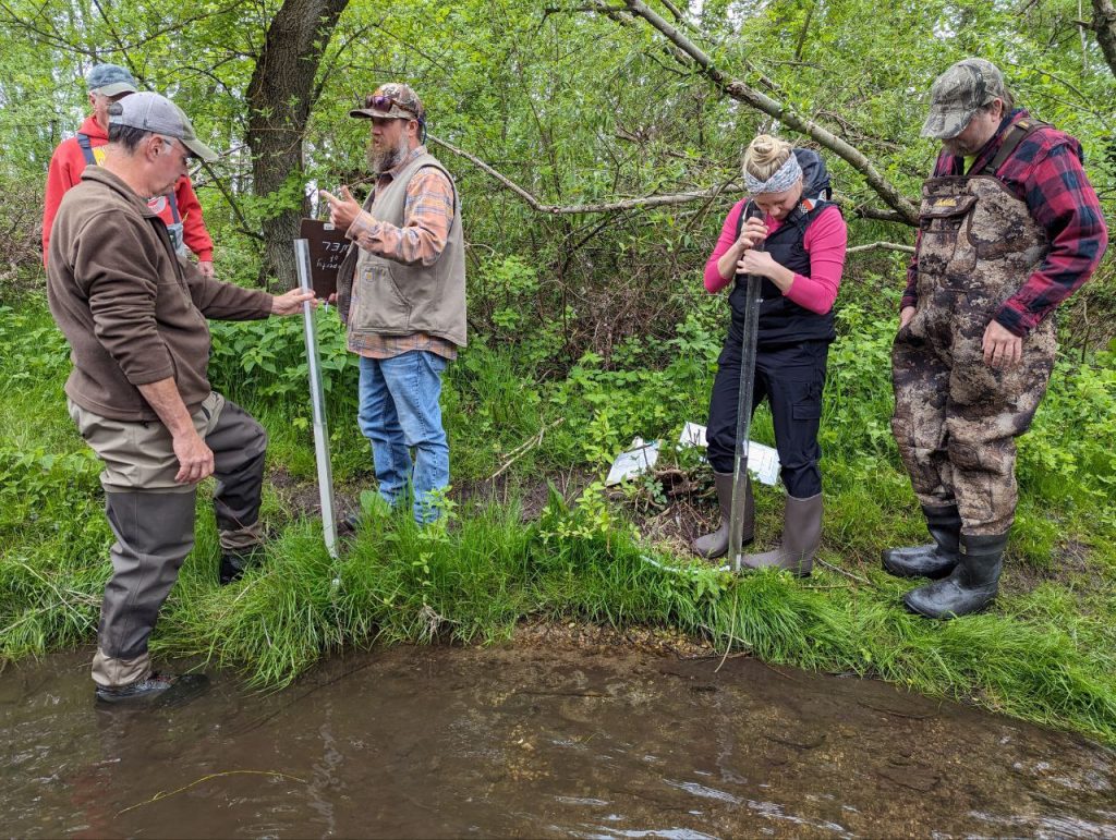A group of five volunteers stand on a stream bank. Two of the volunteers are holding transparency tubes. There is foliage in the background.