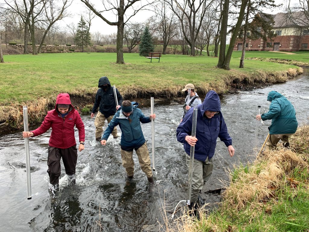 A group of volunteers holding transparency tubes walk through a stream. It is an overcast day. 