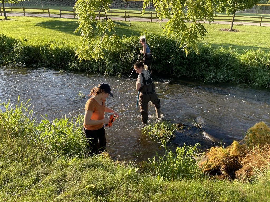 Three volunteers measuring the width of a stream on a sunny day.