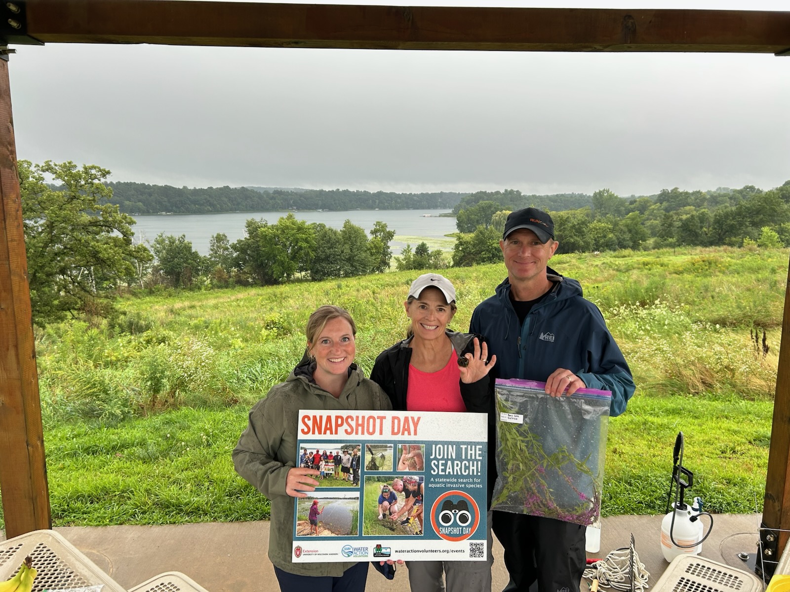 Three volunteers stand under an awning holding a poster that reads "Snapshot Day". In the background is a lake. 