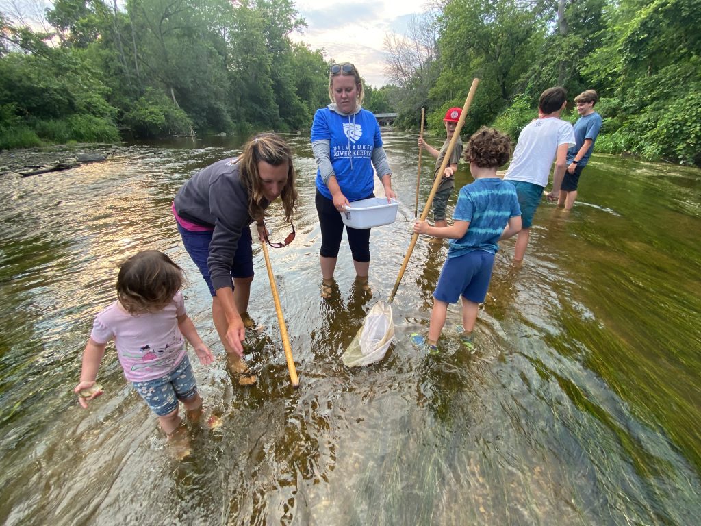 A group of adults and kids stand in a shallow river collecting macroinvertebrates with nets.