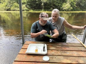 Two volunteers standing a river. They have a tray of dissolved oxygen equipment and are pointing at a piece of paper