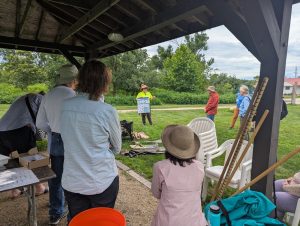 A group of people sit under an awning while an instructor teaches about macroinvertebrates. The instructor is holding a poster with macroinvertebrates.