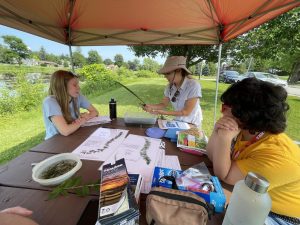 Three people sit at a table in a park alongside a river and learn how to identify plants.