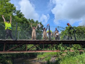 Six volunteers stand on a bridge over a creek. It is a sunny day with a blue sky. 